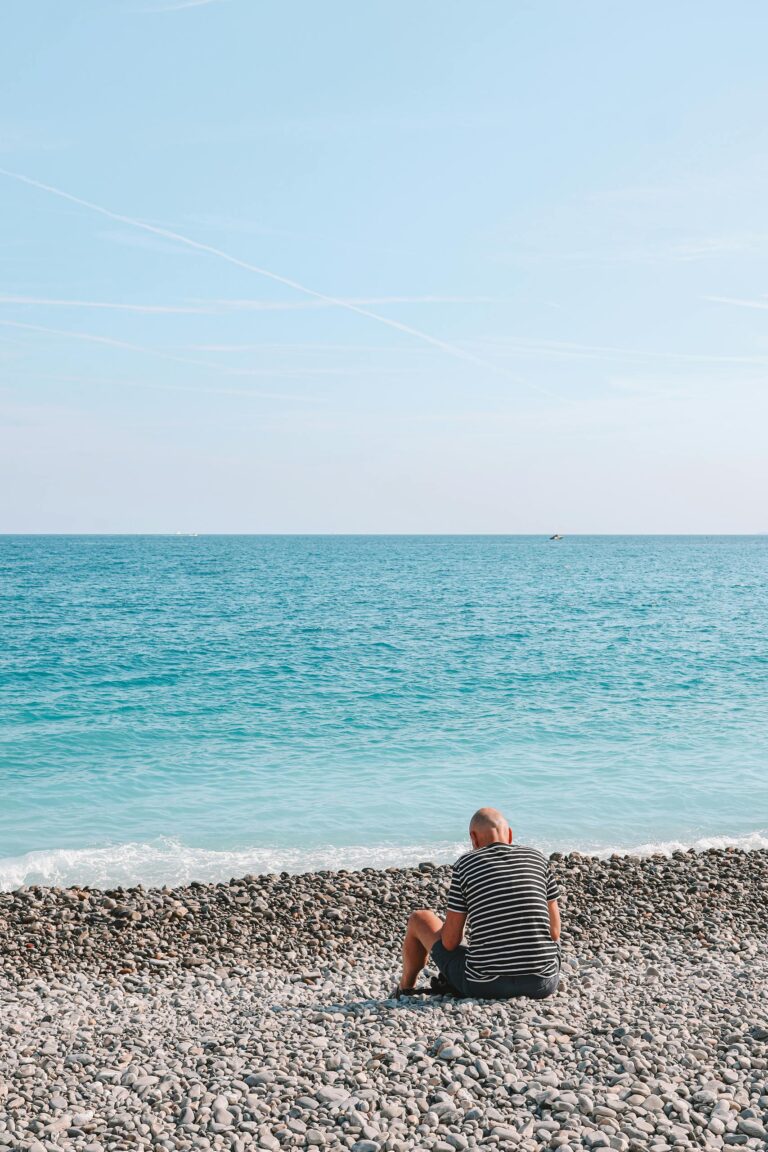 A solitary man sits on a pebble beach in Nice, France, gazing at the clear blue sea under a bright sky.